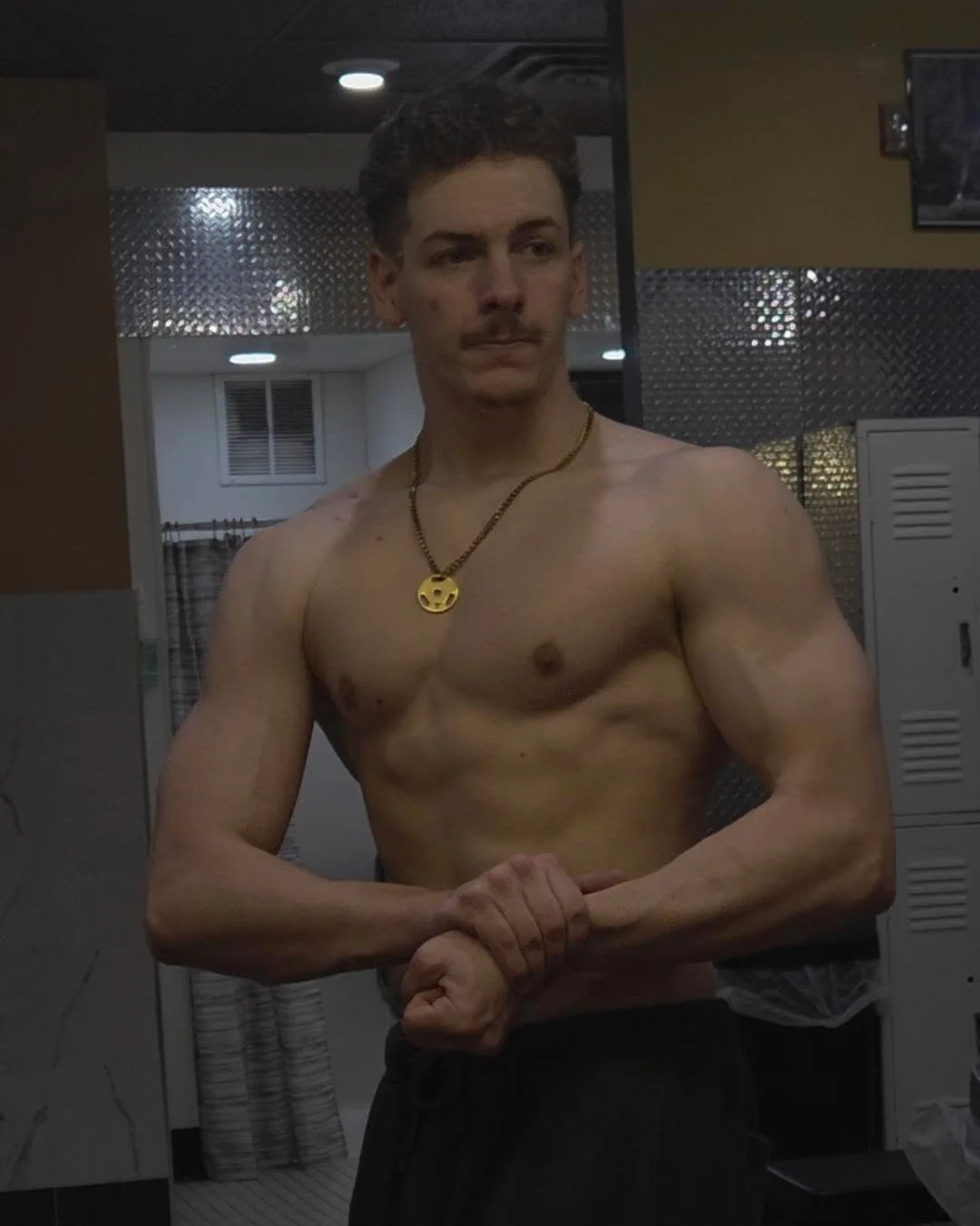 Sam in a gym locker room, shirtless, holding a front-arm pose