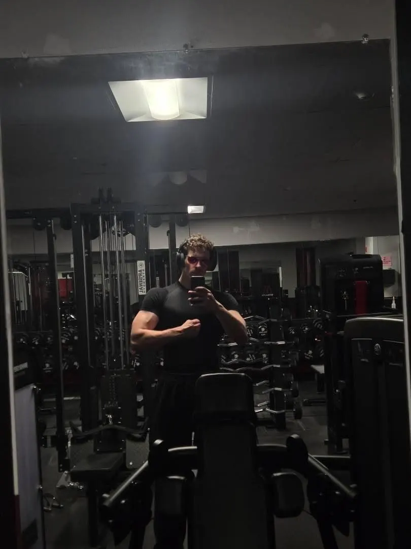 Sam in a dark gym at night, headphones on, flexing a bicep