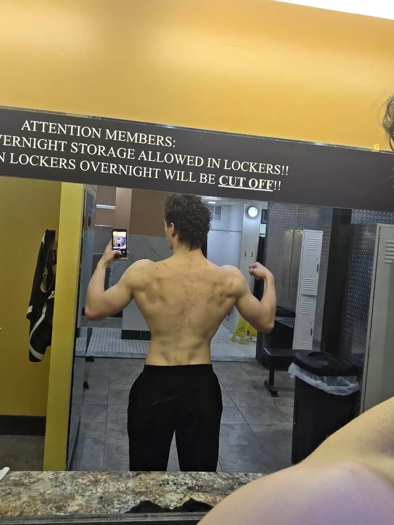 Sam holding a back double biceps pose in a locker room mirror